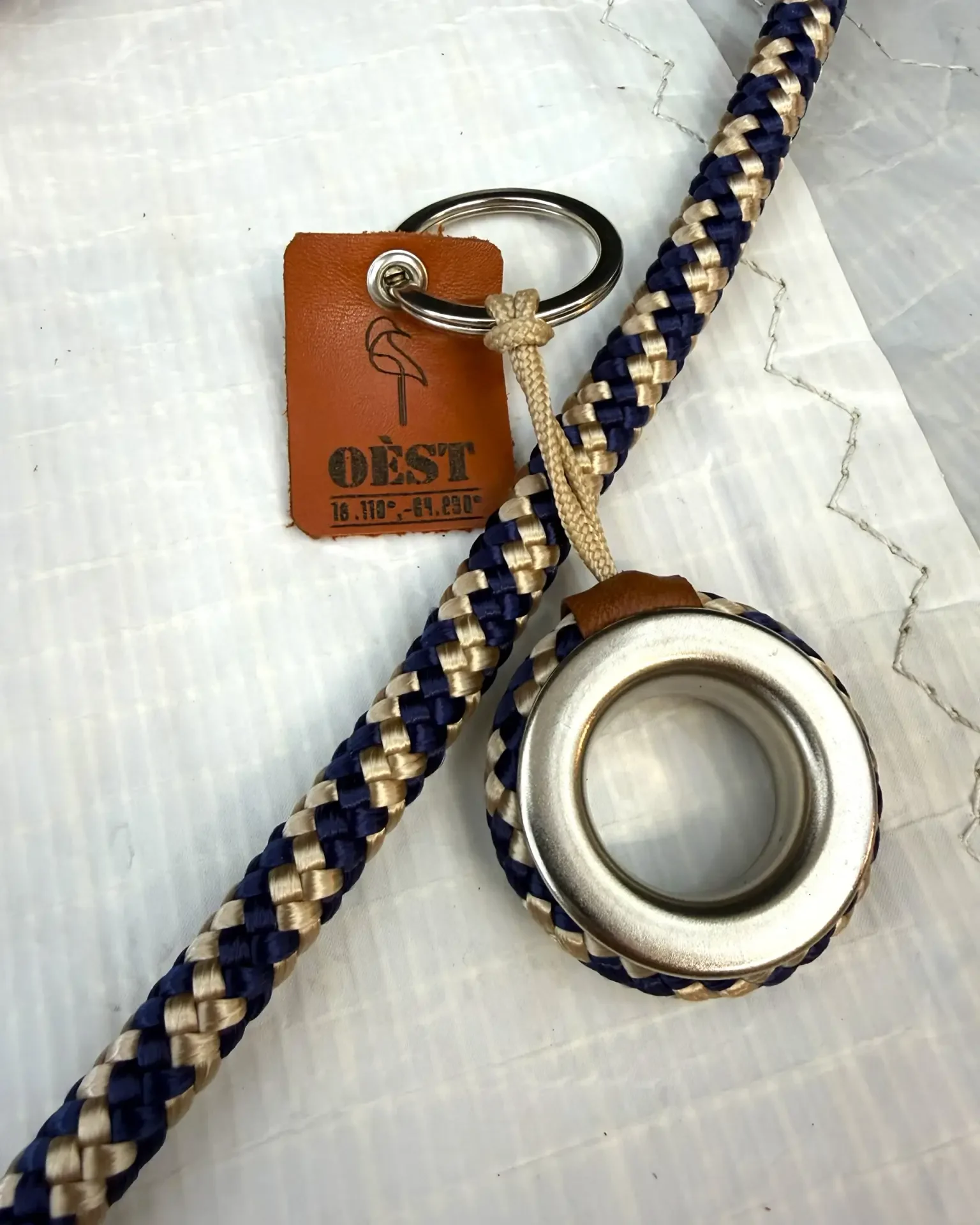Hossegor keyring in navy and beige braided marine rope around a metal eyelet, OÈST tag, lying on a sail